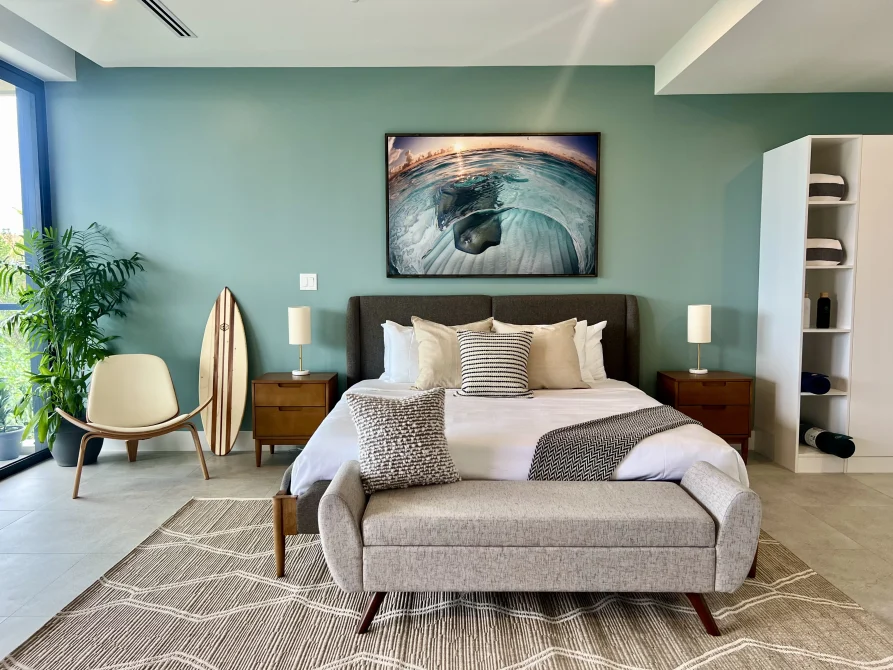 A modern studio suite at Vida Cayman with a king bed, teal accent wall, ocean-themed artwork, surfboard decor, and minimalist furnishings.