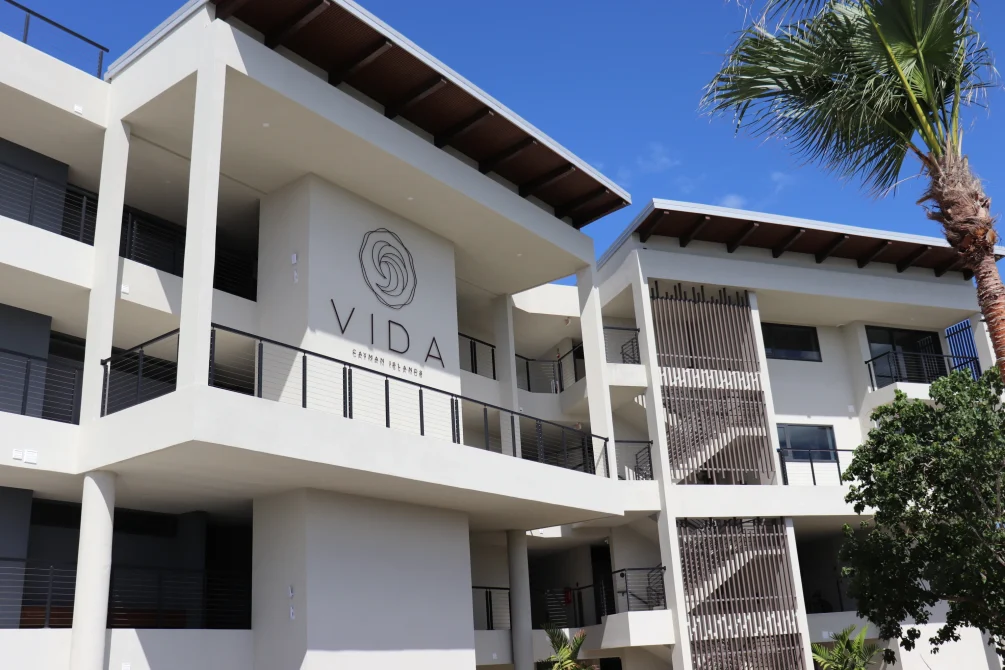 The modern white exterior of Vida Cayman with balconies, palm trees, and the VIDA Cayman Islands logo under a bright blue sky.