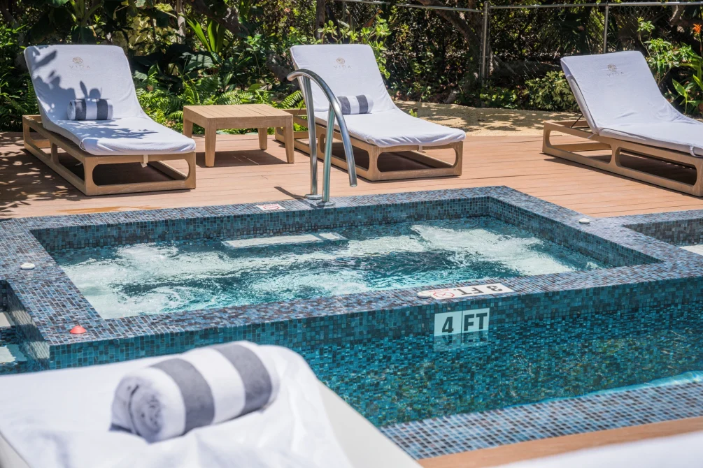 A steaming hot tub surrounded by white loungers, rolled towels, and tropical plants at Vida Cayman's wellness pool area.