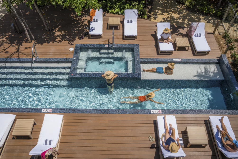 An aerial view of Vida Cayman's pool deck with lap pool, hot tub, loungers, and guests relaxing in the Caribbean sun.