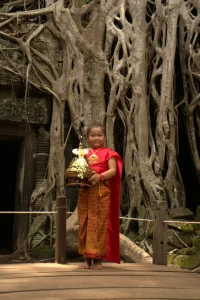 Young Cambodian girl in traditional red and gold silk clothing holding a golden Buddhist offering, standing in front of ancient temple ruins with massive tree roots.