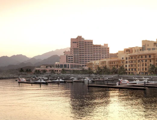 Fairmont Fujairah Beach Resort exterior at sunrise, with terracotta and cream buildings, marina with boats, and dramatic mountain backdrop.