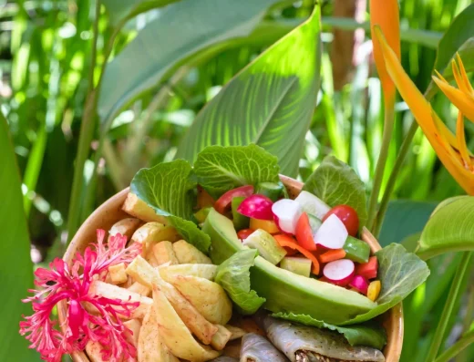A hand holds a colorful bowl of fresh salad, fries, and wraps garnished with tropical flowers, set against lush green foliage and orange heliconia blooms.