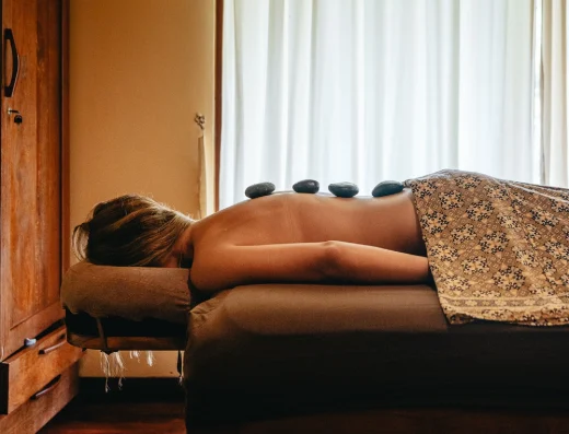 A woman receives a hot stone massage on a spa bed with warm earth tones and soft natural light streaming through curtains.