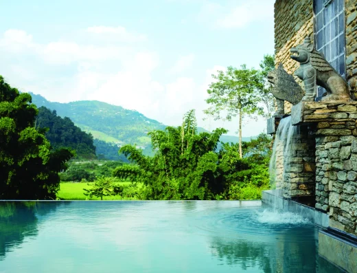 An infinity pool with turquoise water overlooking lush green Himalayan valleys, forested mountains, and a stone water feature with decorative sculpture.