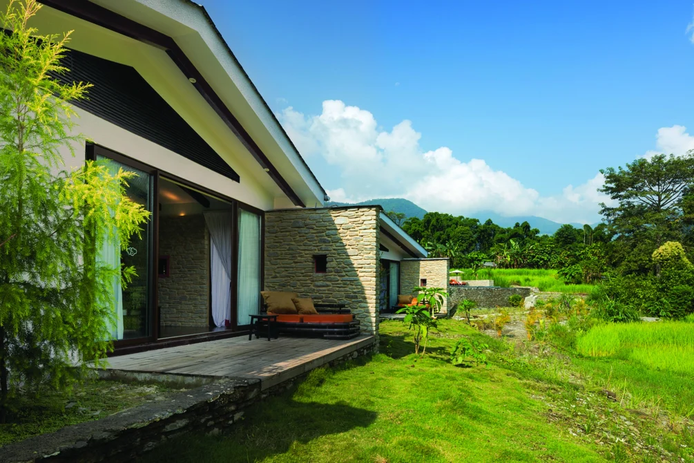 A modern villa with stone and white walls, wooden deck, and large windows overlooking rice paddies and Himalayan mountains under clear blue sky.