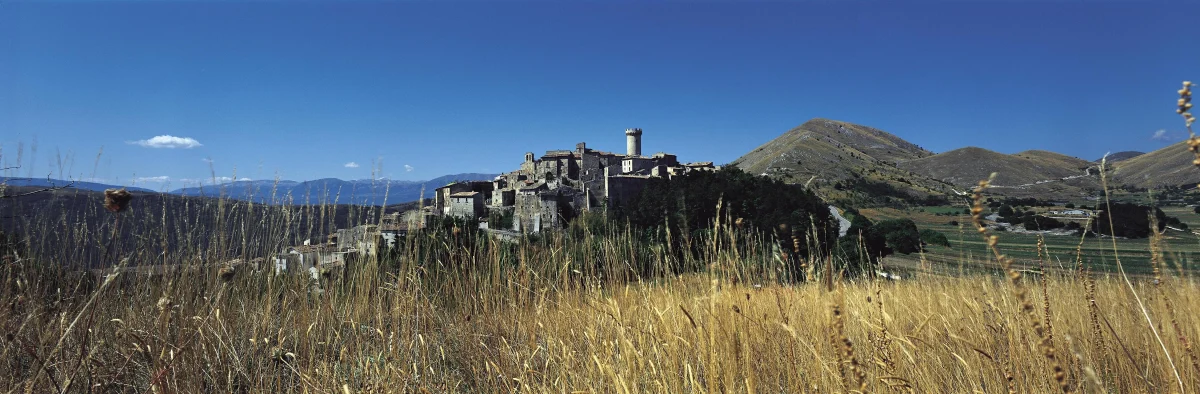 Sextantio Santo Stefano di Sessanio- A medieval hilltop village with stone towers and buildings surrounded by golden wheat fields, mountains, and blue sky in Abruzzo, Italy.