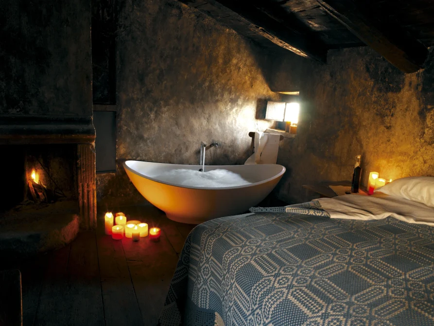 A cave bedroom with a sculptural golden soaking tub, fireplace, candlelit ambiance, and a bed with patterned blanket carved into ancient stone walls.