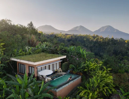 A villa with an infinity pool sits above lush tropical forest, with misty mountains in the distance under a clear sky.