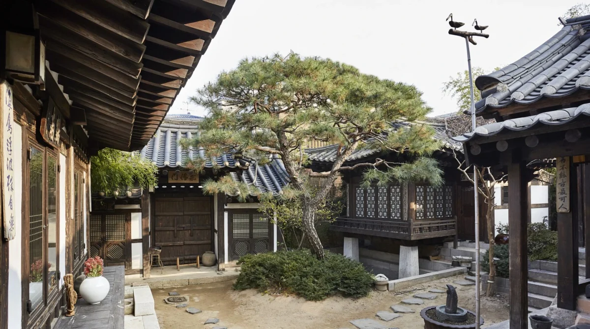 A traditional Korean hanok courtyard with curved tile roofs, wooden pavilions, ancient pine tree, and stone pathways in Seoul's Bukchon village.