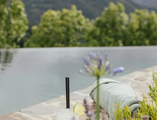 A cocktail with lime sits beside a towel on a stone pool deck overlooking green Asturian mountains and a calm infinity pool at Solo Palacio.