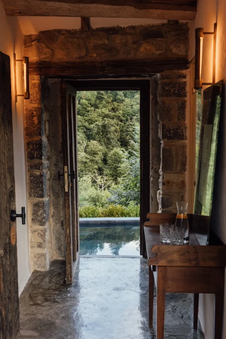 A stone doorway opening onto a reflecting pool and lush Asturian forest, with warm interior lighting and a wooden table set with glasses.