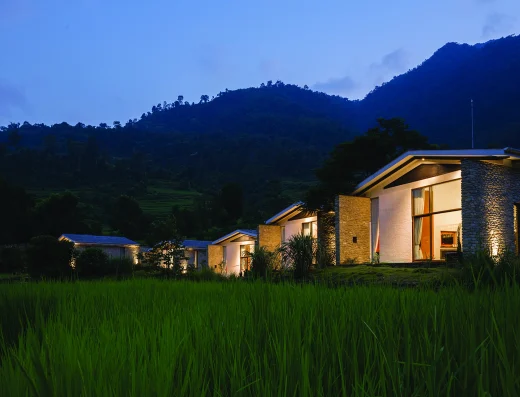 Modern stone villas with warm lighting at dusk, set among rice paddies with forested Himalayan mountains rising in the background.