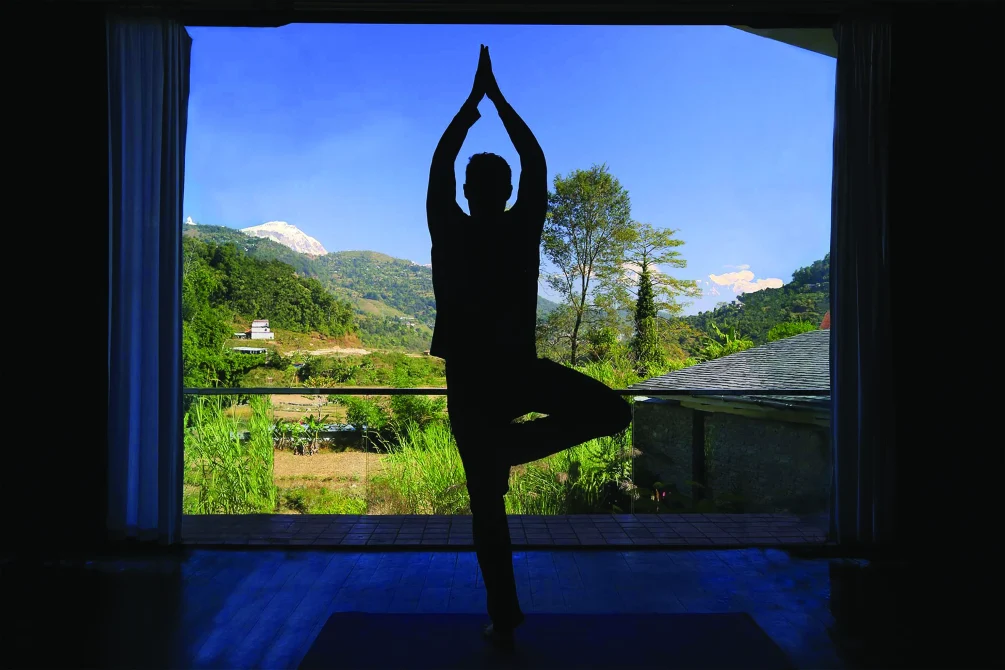 A silhouetted yoga practitioner in tree pose inside a studio with floor-to-ceiling windows framing snow-capped Annapurna mountains and lush Himalayan valleys.