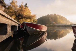 Red and black wooden boats moored at a dock on a misty Alpine lake, with golden autumn trees and forested mountains reflected in still water.