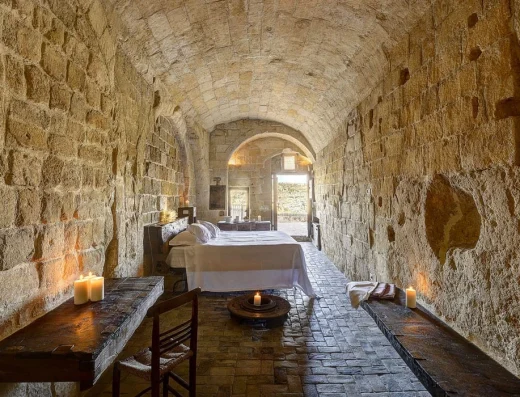 A candlelit cave-suite bedroom in Matera with arched stone ceiling, rustic wooden furniture, and a bed set deep inside ancient limestone walls.