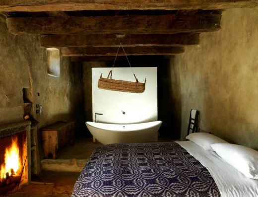 A cave suite bedroom with a fireplace, exposed wooden beams, a bed with patterned blanket, and a freestanding bathtub set into the stone alcove.