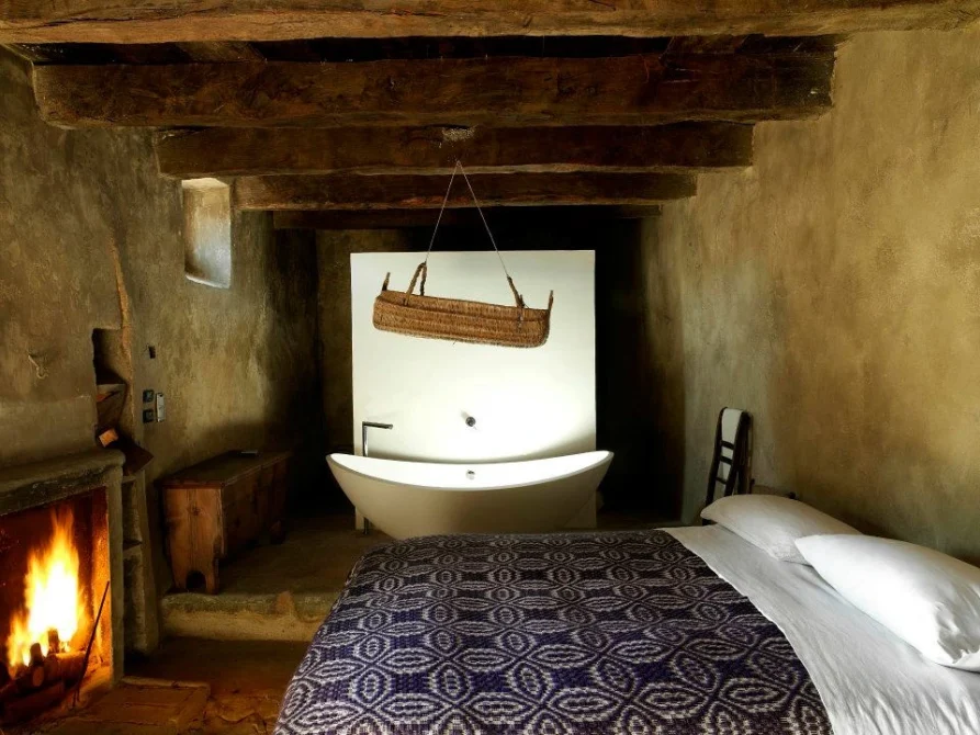 A cave suite bedroom with a fireplace, exposed wooden beams, a bed with patterned blanket, and a freestanding bathtub set into the stone alcove.