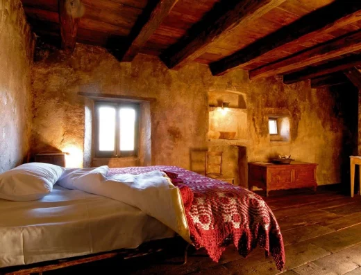 A rustic cave bedroom with exposed wooden beams, terracotta walls, a bed with red woven blanket, and warm natural light from a small window.