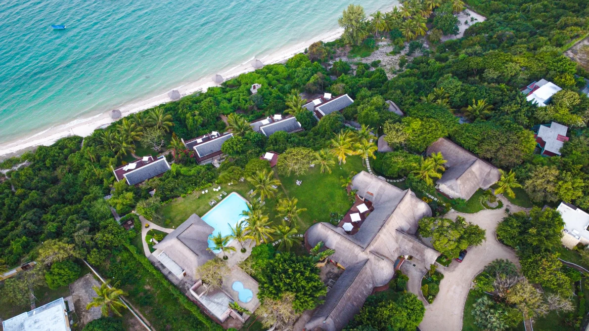 Aerial view of a beachfront resort with a pool, thatched-roof buildings, and lush tropical gardens beside the ocean.