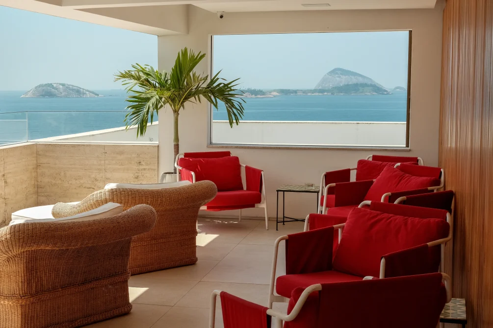 Ocean-view lounge with red armchairs and wicker seating, overlooking the sea and distant islands.