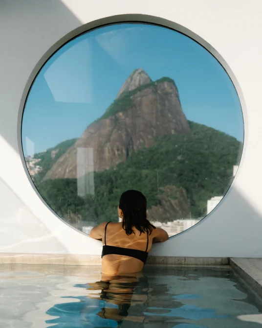 Guest relaxing in a pool beneath a circular window framing a mountain view.