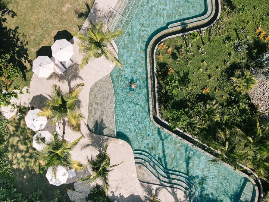 Aerial view of a curving resort pool surrounded by palm trees, lush gardens, and white sun umbrellas.