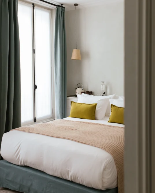 Bright, minimalist bedroom with a double bed, white linens, mustard cushions, and a soft blue curtain by a tall window. Hotel HOY Paris