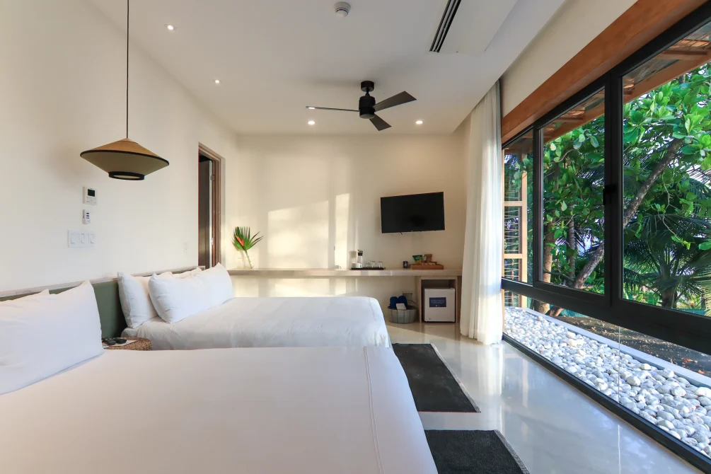 Bright modern bedroom with two beds, white linens, and large windows looking out to tropical trees.