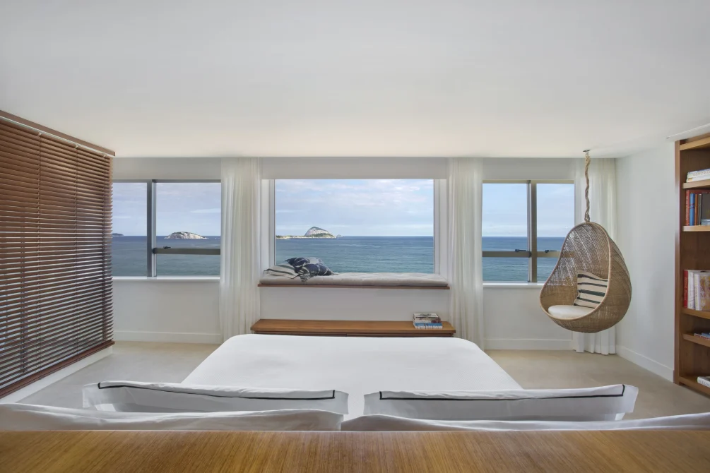 Ocean-view bedroom with wide windows, window seat, and hanging wicker chair overlooking the sea.